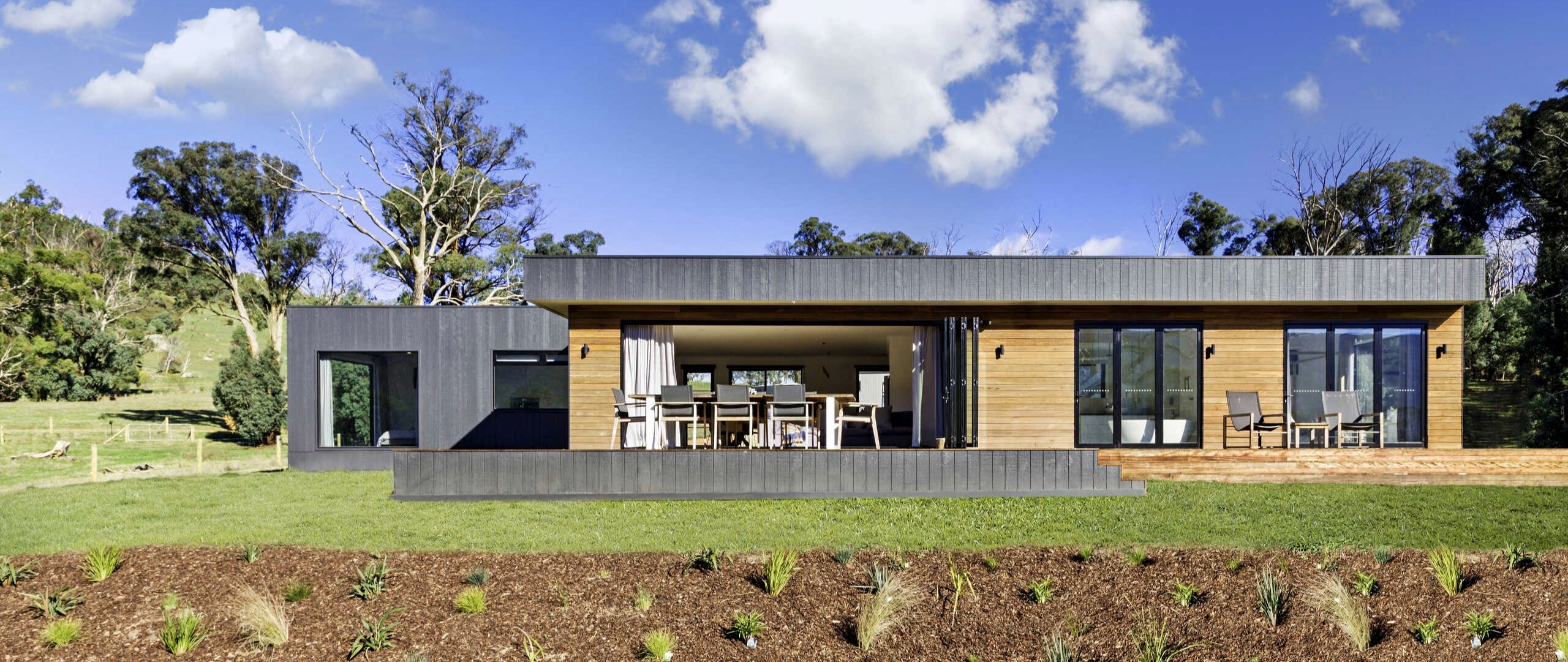 Exterior of a three bedroom modular home by Anchor Homes in Buxton in the Yarra Valley Victoria, featuring dual cladding with dark Nailstrip and warm timber panels, a flat roof design, open bifold doors to an alfresco dining area, and a freshly landscaped garden bed with rural views beyond