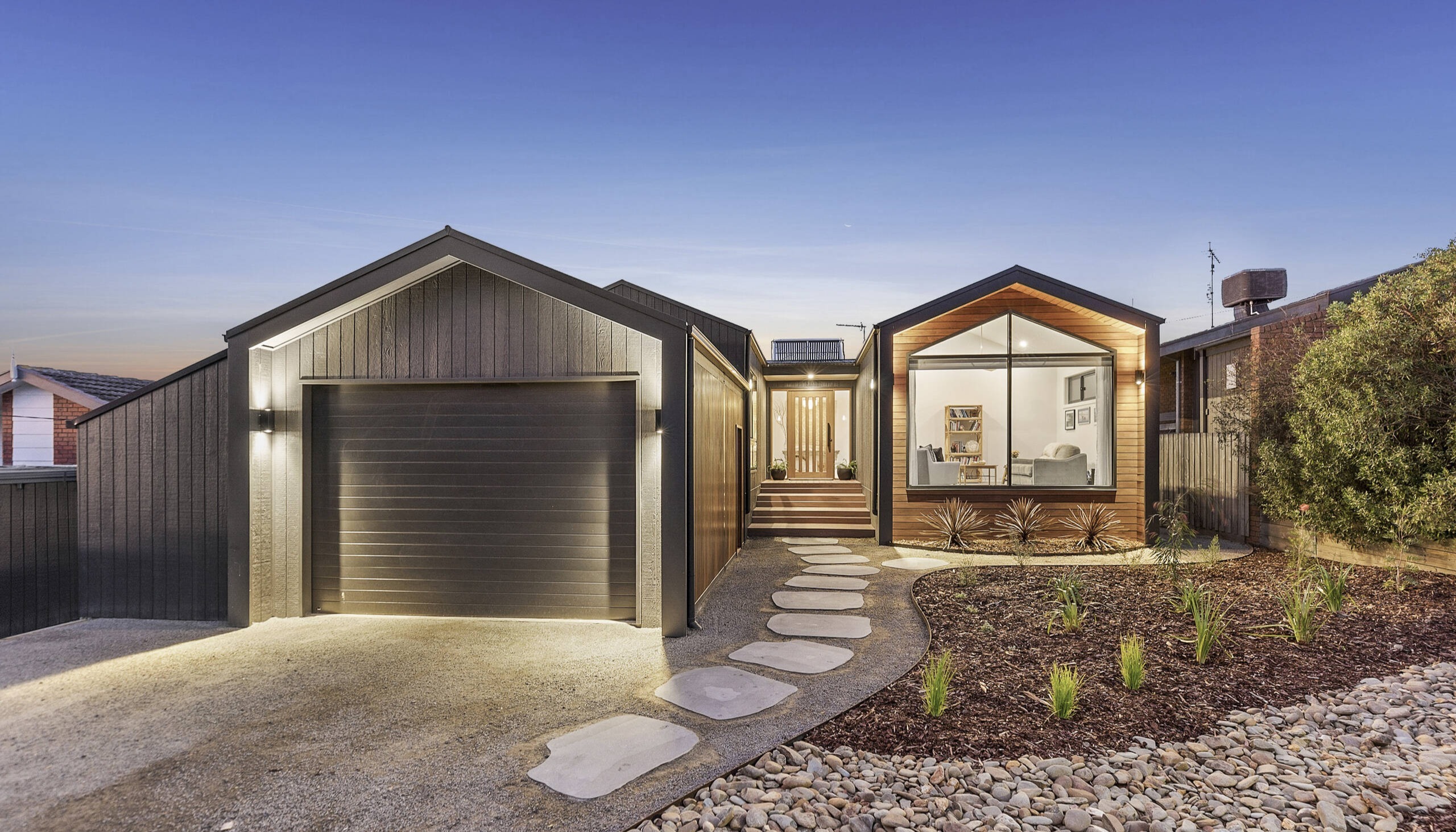 Front facade of the Portsea 16 modular home by Anchor Homes in Clifton Springs on the Bellarine Peninsula Victoria, photographed at dusk with warm interior lighting, dark and timber dual cladding, a double garage, modern gable roofline, stepping stone path, and low maintenance coastal landscaping