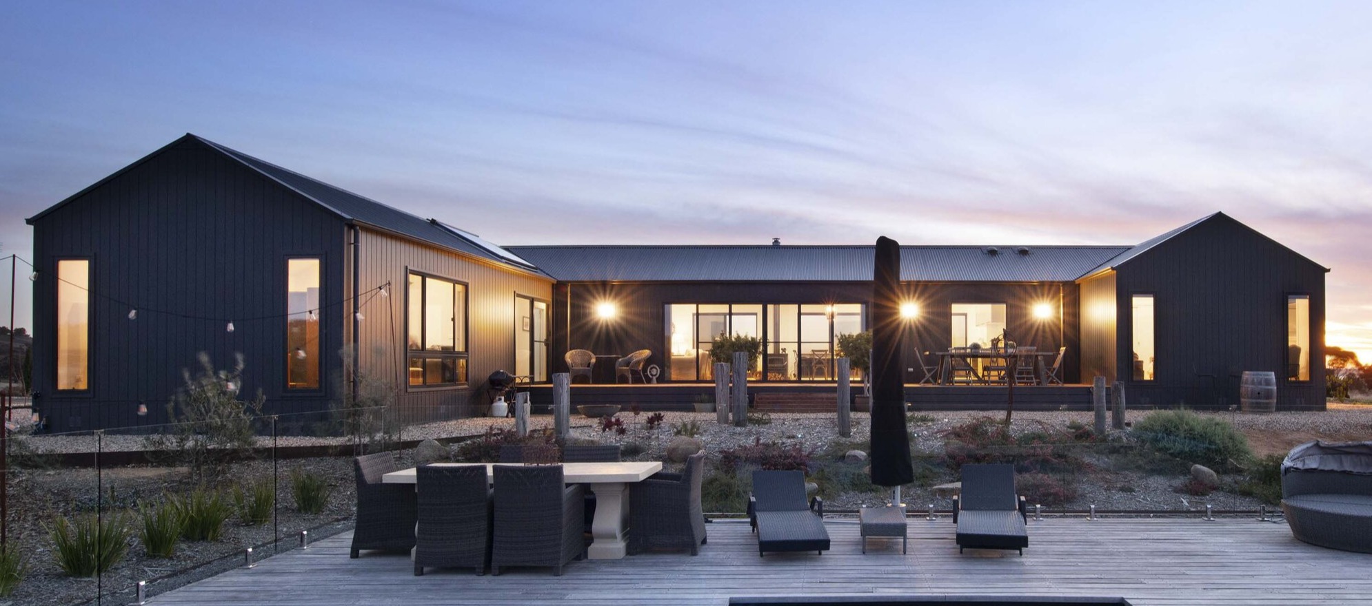 Three bedroom Newhaven 21 prefab home by Anchor Homes in Tarrengower Victoria, photographed at dusk with a illuminated swimming pool and timber deck in the foreground, outdoor dining and lounge furniture, and the dark Nailstrip clad facade glowing warmly against a sunset sky